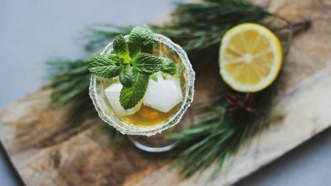 Ein Cocktail mit frischer Minze und Eiswürfeln steht auf einem Holzbrett, umgeben von Tannenzweigen und einer Zitronenscheibe.