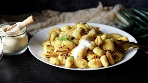 Gnocchi liegen gebraten auf einem Teller, garniert mit Crème Fraîche. Daneben steht ein Glas mit einem Holzlöffel auf einem dunklen Tisch.