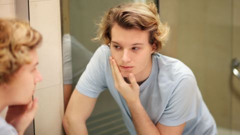 Ein junger Mann betrachtet besorgt sein Gesicht im Spiegel, während er seine Haut mit einer Hand berührt.