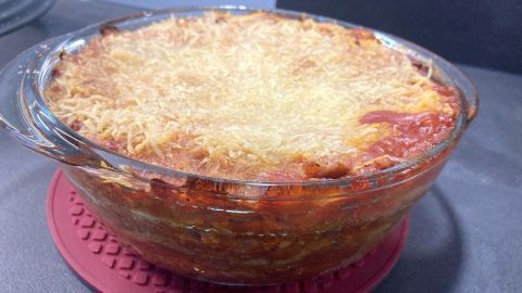Eine goldbraune vegane Lasagne in einer Glasform steht auf einem roten Untersetzer, umgeben von dunklem Küchenuntergrund.