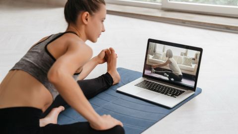 Eine Person macht Yoga-Übungen auf einer Matte, während sie ein YouTube-Video auf einem Laptop verfolgt.