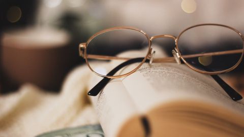 Eine Brille liegt auf einem aufgeschlagenen Buch, während eine Tasse Kaffee im Hintergrund für eine gemütliche Atmosphäre sorgt.