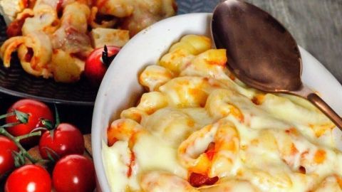 Ein Auflauf mit Käse und Nudeln wird serviert, umgeben von frischen Tomaten auf einem rustikalen Tisch.