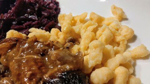 Zarte Spätzle liegen neben einer Fleischscheibe in einer Soße und rotem Kraut auf einem weißen Teller.