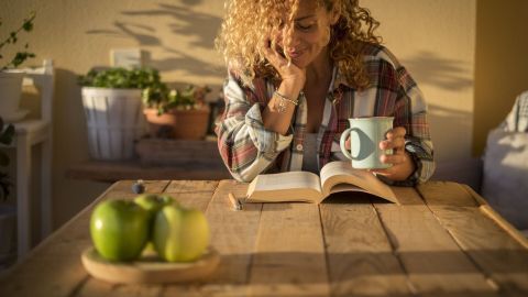 Eine nachdenkliche Frau sitzt an einem Holztisch, liest ein Buch und hält eine Tasse, während Äpfel daneben stehen.