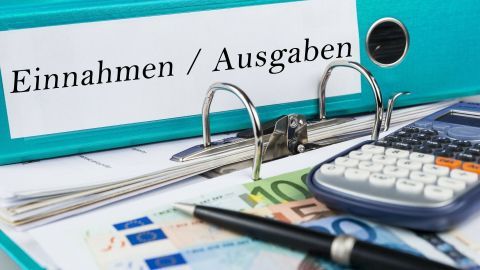 Ein Ordner mit der Beschriftung "Einnahmen / Ausgaben" liegt auf einem Tisch, umgeben von Geldscheinen und einem Taschenrechner.