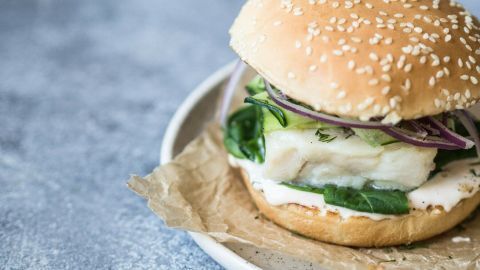 Ein weicher Burgerbrötchen mit frischem Fisch, Spinat, Gurken und Zwiebeln auf einem Teller mit braunem Papier.