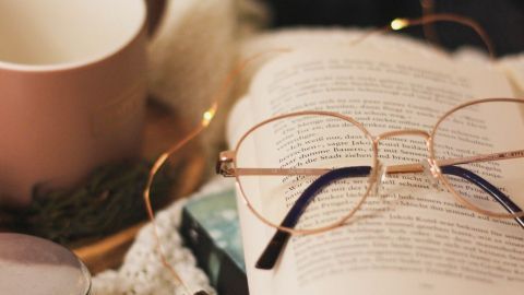 Ein offenes Buch liegt auf einem gestrickten Tuch, während eine Brille daneben liegt; eine Tasse und Dekoration schaffen eine gemütliche Atmosphäre.