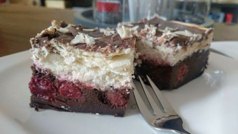 Ein Stück Schwarzwälder Käsekuchen liegt auf einem weißen Teller, garniert mit Schokolade und Kirschen, neben einer Gabel.