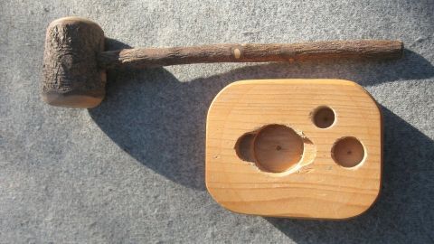 Ein Holzhammer liegt neben einem aus Holz gefertigten Nussknacker, der runde Vertiefungen zum Knacken hat, auf einem grauen Untergrund.
