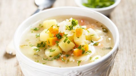 Eine weiße Schüssel mit Möhren-Senf-Suppe, garniert mit Gemüsewürfeln und Kräutern, steht auf einem Holztisch.