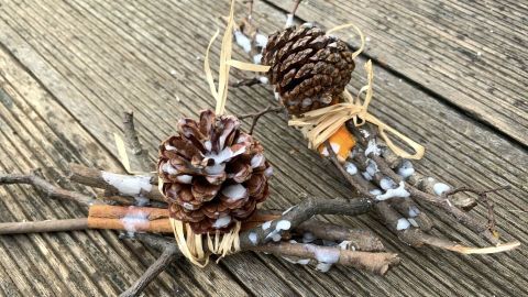 Zwei handgemachte Kaminanzünder aus Tannenzapfen und Ästen liegen auf einem Holzbrett. Sie sind mit dünnen Bändern gebunden.