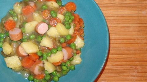 Erbseneintopf mit Kartoffeln, Karotten und Wurststücken in einer blauen Schüssel auf einem Holzuntergrund serviert.