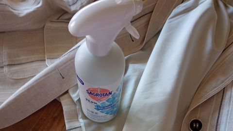 Ein Sprayflasche steht auf einem Holzfußboden neben einer beigen Jacke, um Zigarettengeruch aus Klamotten zu entfernen.