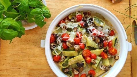 Ein vegetarischer Maultaschenauflauf wird in einer weißen Schüssel mit buntem Gemüse und Käse auf einem Holzbrett serviert.