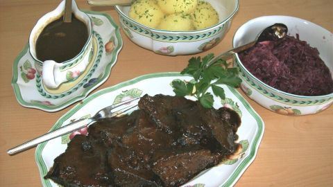 Ein saftiger Sauerbraten liegt auf einem Teller, umgeben von roten Kohl, Kartoffeln und Soße in eleganten Schüsseln.