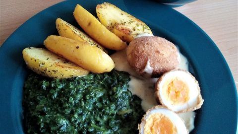 Frittierte Eier liegen neben Rahmspinat und knusprigen Kartoffeln auf einem blauen Teller, während eine Schüssel Suppe daneben steht.
