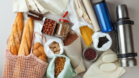 Ein Netzbeutel enthält frisches Brot, während diverse nachhaltige Lebensmittel und eine Thermosflasche auf einem Tisch liegen.
