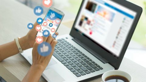 Eine Person hält ein Smartphone, während sie mit einem Laptop arbeitet. Soziale Medien-Symbole erscheinen um das Handy.