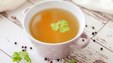 Eine schlichte Tasse mit klarer, goldener Brühe und frischen Kräutern befindet sich auf einem rustikalen Holztisch.