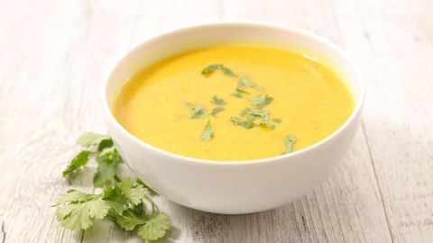 Eine cremige Tomatensuppe steht in einer weißen Schüssel, garniert mit frischen Kräutern, auf einem hellen Holztisch.