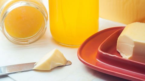 Ein Glas mit gelber Flüssigkeit steht neben einem Stück Butter und einem weiteren Glas auf einem Tisch.