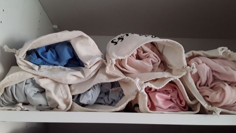 Gerollte Spannbettlaken liegen ordentlich in Stoffbeuteln auf einem Regal, gut organisiert und sichtbar gelagert.