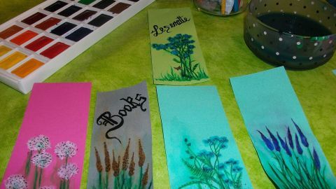 Bunte Lesezeichen liegen auf einem Tisch, umgeben von Wasserfarben und einem Glas mit Wasser, während gemalt wird.