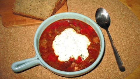 Eine Schüssel mit scharfer Gulaschsuppe steht neben geröstetem Brot auf einem Tisch, während ein Löffel bereitliegt.