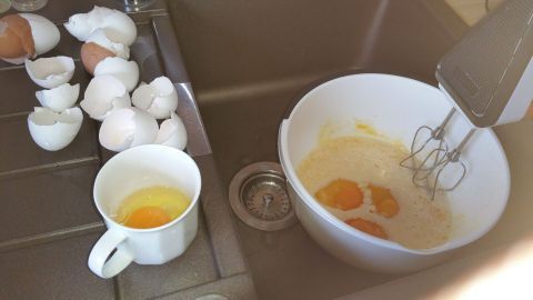 In einer Küchenszene werden Eier einzeln aufgeschlagen, während einige Schalen daneben liegen, und ein Mixer in einer Schüssel steht.