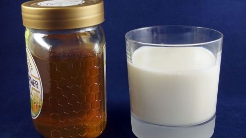 Ein Glas frische Milch steht neben einem Honigglas, beide sind auf einem blauen Hintergrund platziert, ideal für Aknebehandlungen.