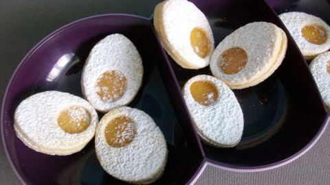 Ostereier-Plätzchen aus Mürbeteig liegen auf einem lila Teller, gefüllt mit Aprikosenkonfitüre und Puderzucker.