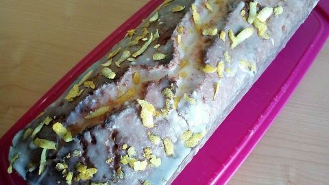 Ein Zitruskuchen liegt auf einem rosa Behälter, glasiert und mit orangefarbener Schale dekoriert, auf einem hellen Tisch.