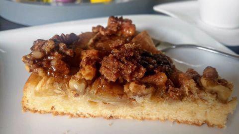Ein Stück Apfelkuchen mit karamellisierten Walnüssen liegt auf einem weißen Teller, umgeben von bunten Dekorationen im Hintergrund.