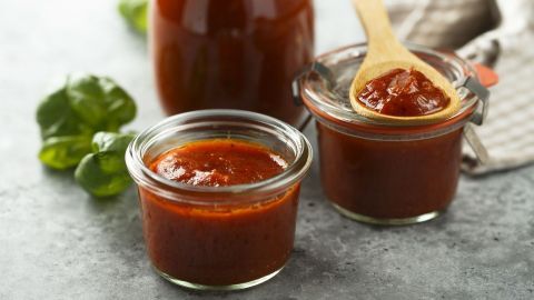 Zwei Gläser mit selbstgemachtem Tomatenketchup stehen auf einem grauen Tisch, ein Löffel hebt die würzige Soße heraus.