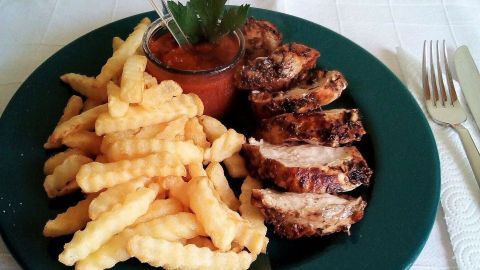Gegrillte Hähnchenfilets liegen neben knusprigen Pommes auf einem grünen Teller und werden von einer Tomatensauce begleitet.