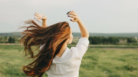 Eine Frau schwingt ihr langes, brünettes Haar in einer grünen, offenen Landschaft, während sie entspannend in die Ferne blickt.