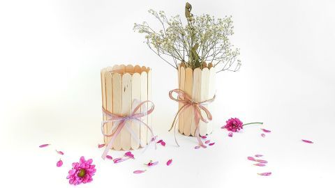 Zwei DIY Upcycling-Vasen aus Holzstäbchen halten Trockenblumen, umgeben von Blütenblättern auf weißem Hintergrund.