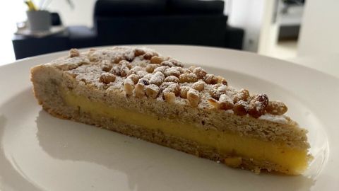 Ein Stück Torta della Nonna liegt auf einem weißen Teller, umgeben von einem modernen Wohnraum mit einem Sofa im Hintergrund.