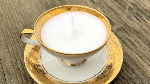 In einer eleganten, goldverzierten Tasse steht eine weiße Kerze auf einem passenden Unterteller auf einer rustikalen Holzoberfläche.