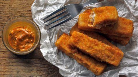 Fischstäbchen liegen auf zerknülltem Papier, während eine Gabel daneben hastig das scharfe Curry-Dip probiert.
