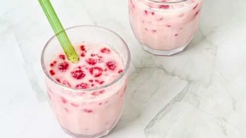 Zwei Gläser mit einem rosa Getränk, verziert mit Himbeeren, stehen auf einer hellen, marmorierten Oberfläche.