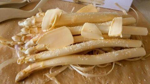 Weißer Spargel liegt auf einem Holzbrett, vorbereitet mit einem Sparschäler und einem Karton im Hintergrund.