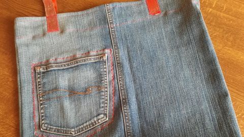 Eine handgenähte Einkaufstasche aus Jeans liegt auf einer Holzoberfläche. Die Tasche hat rot-orangefarbene Träger und eine aufgenähte Rückentasche.