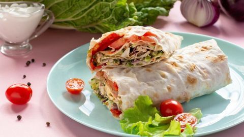 Zwei leckere Wraps mit frischen Tomaten und Salat liegen auf einem Teller, umgeben von Gemüse und einer Schale Joghurt.