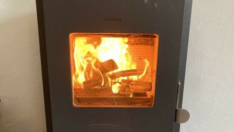 Ein kleiner Kaminofen brennt mit loderndem Feuer und Holzscheiten, gestellt auf einem dunklen Boden in einem gemütlichen Raum.