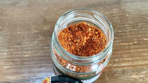 Ein Glas mit Piment d'Espelette ist geöffnet, der Inhalt ist grob gemahlen und auf einem Holztisch platziert.