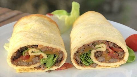 Eine Low Carb Big Mac Rolle liegt auf einem weißen Teller, gefüllt mit Fleisch, Salat und Tomaten, auf einem Holztisch.