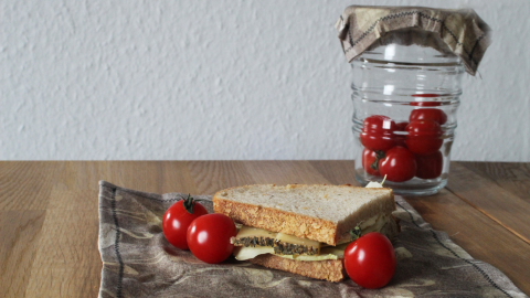 Ein Bienenwachstuch liegt auf einem Holztisch, während ein belegtes Sandwich und frische Tomaten darauf präsentiert werden.