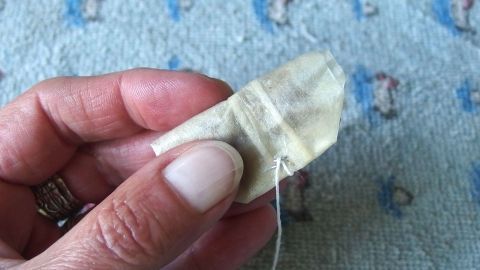 Eine Hand hält einen kleinen Teebeutel, der mit Kamillentee gefüllt ist, auf einem bunten, gemusterten Untergrund.